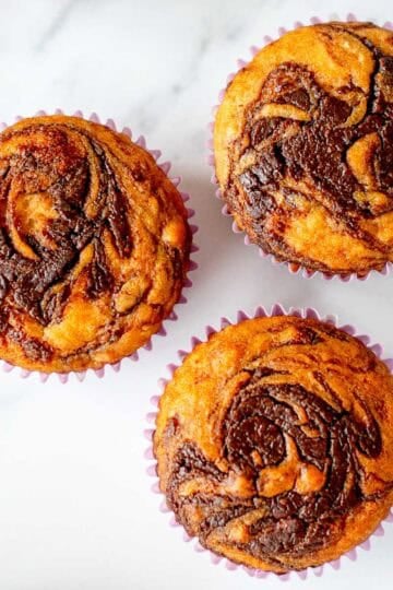 3 Muffins with a Swirl of Nutella on top, sitting on a white marble benchtop.