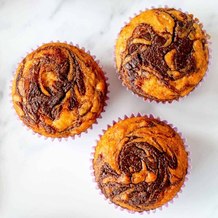 3 Muffins with a Swirl of Nutella on top, sitting on a white marble benchtop.