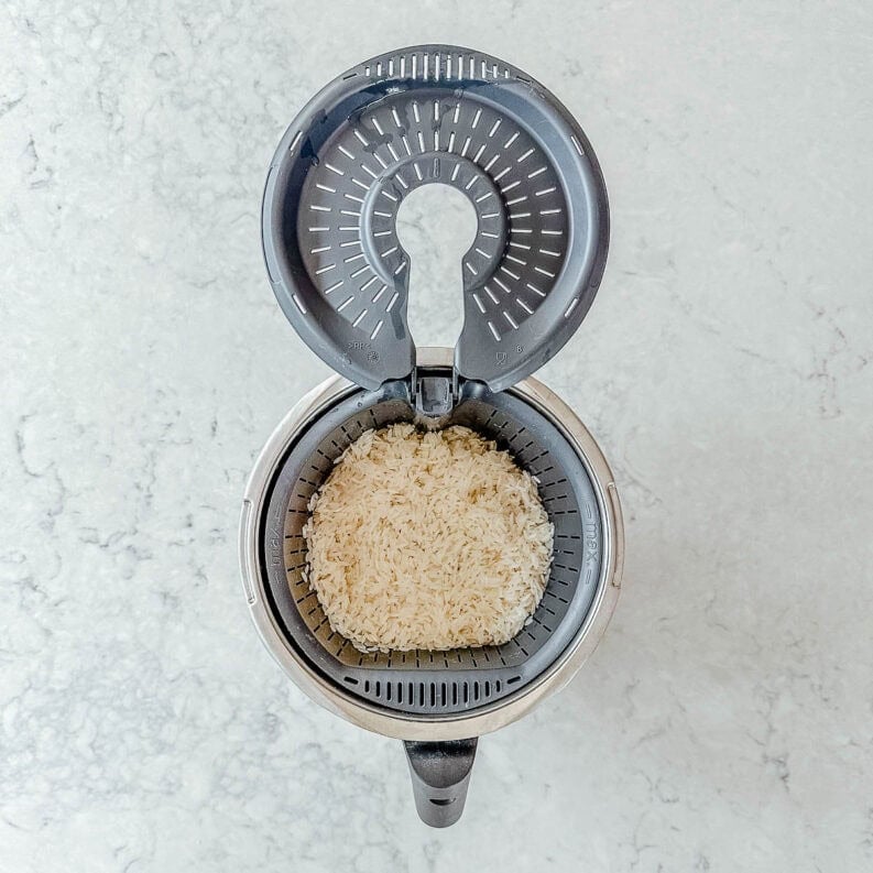 Overhead view of an open thermomix with uncooked white rice inside, on a light-coloured marble worktop.