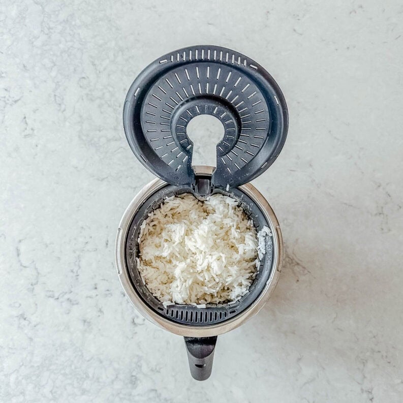Overhead view of an open rice cooker with cooked white rice inside, on a light-coloured marble worktop.