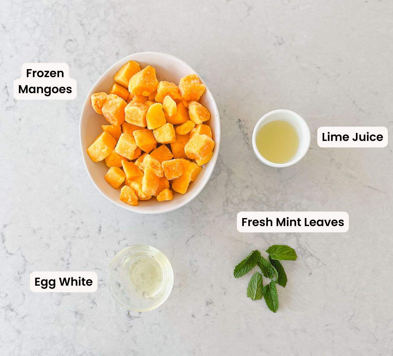 Frozen mango chunks in a white bowl, lime juice in a white bowl, fresh mint leaves and egg white in a clear bowl. all sitting on a marble benchtop.