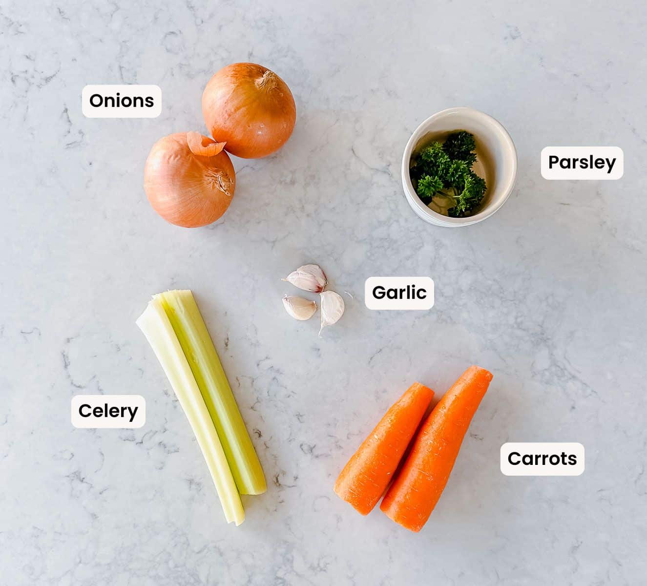 Celery, onion, parsley, garlic and carrot on a marble benchtop.