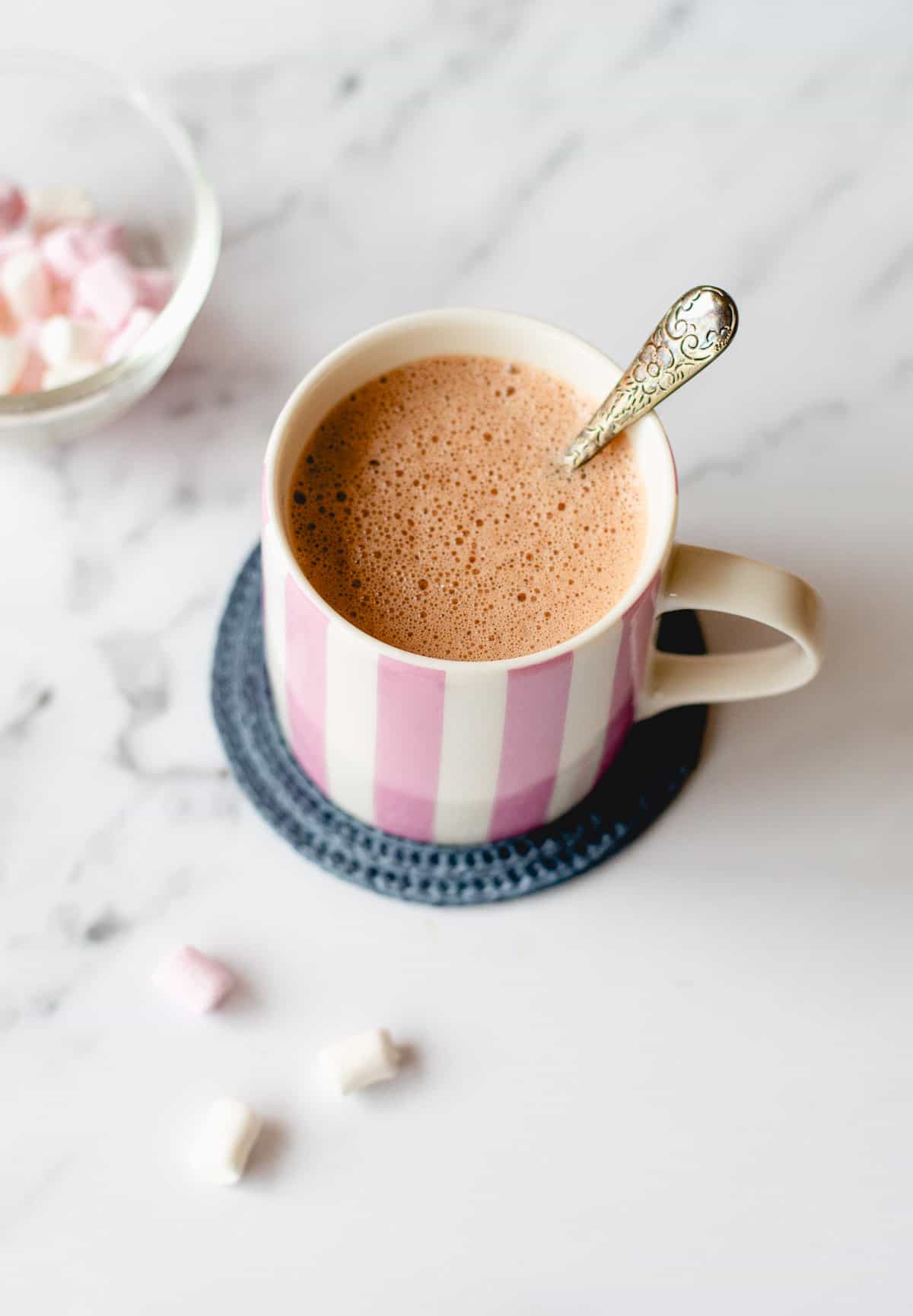 An image of hot chocolate and marshmallows in a pink and white stripy mug on a blue coaster and marble background.