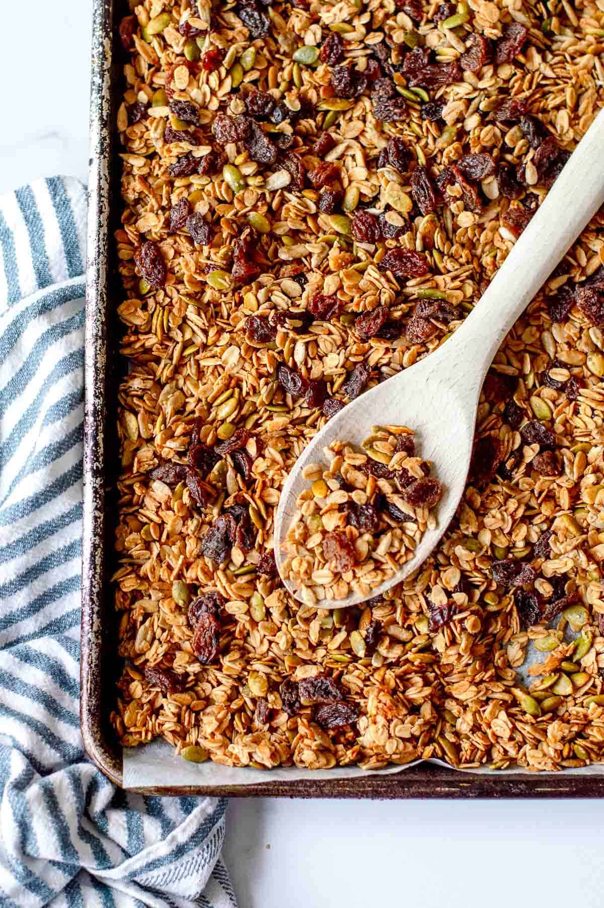 Homemade granola on a baking tray with wooden spoon and blue stripy tea towel.