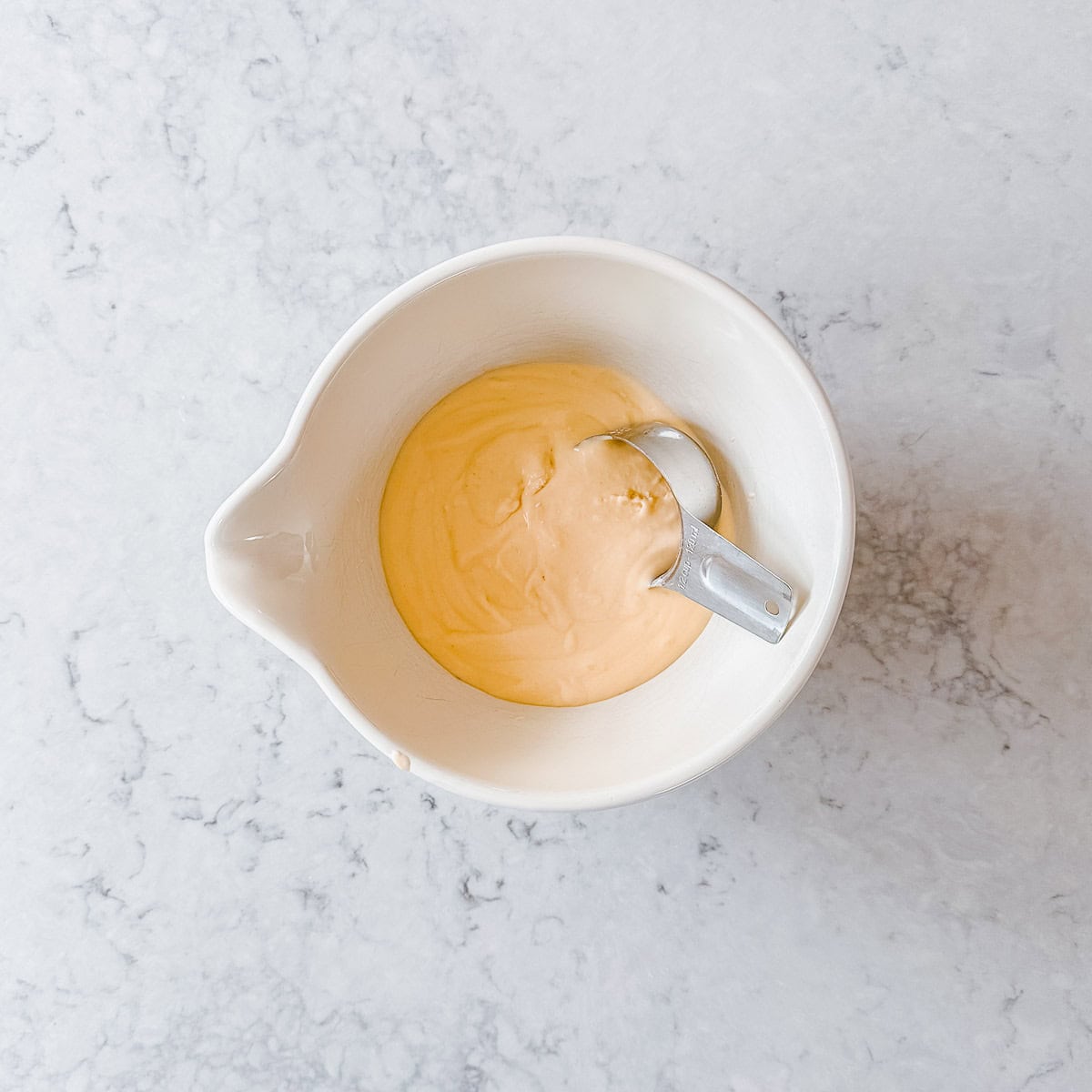 Waffle batter in a cream bowl with metal measuring cup, sitting on a marble benchtop.