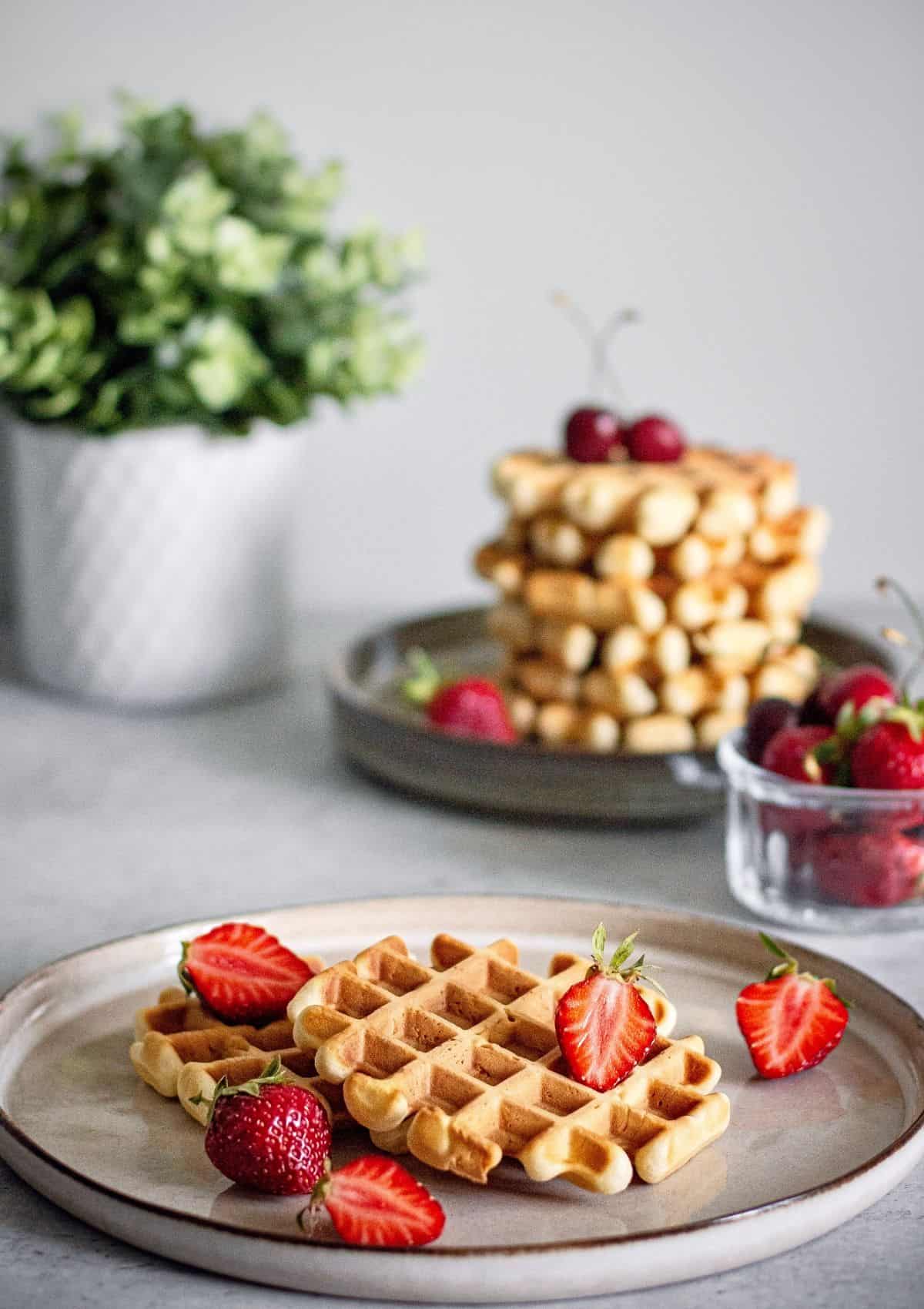 Waffles on a grey plate with strawberries. A waffle stack is seen in the distance.