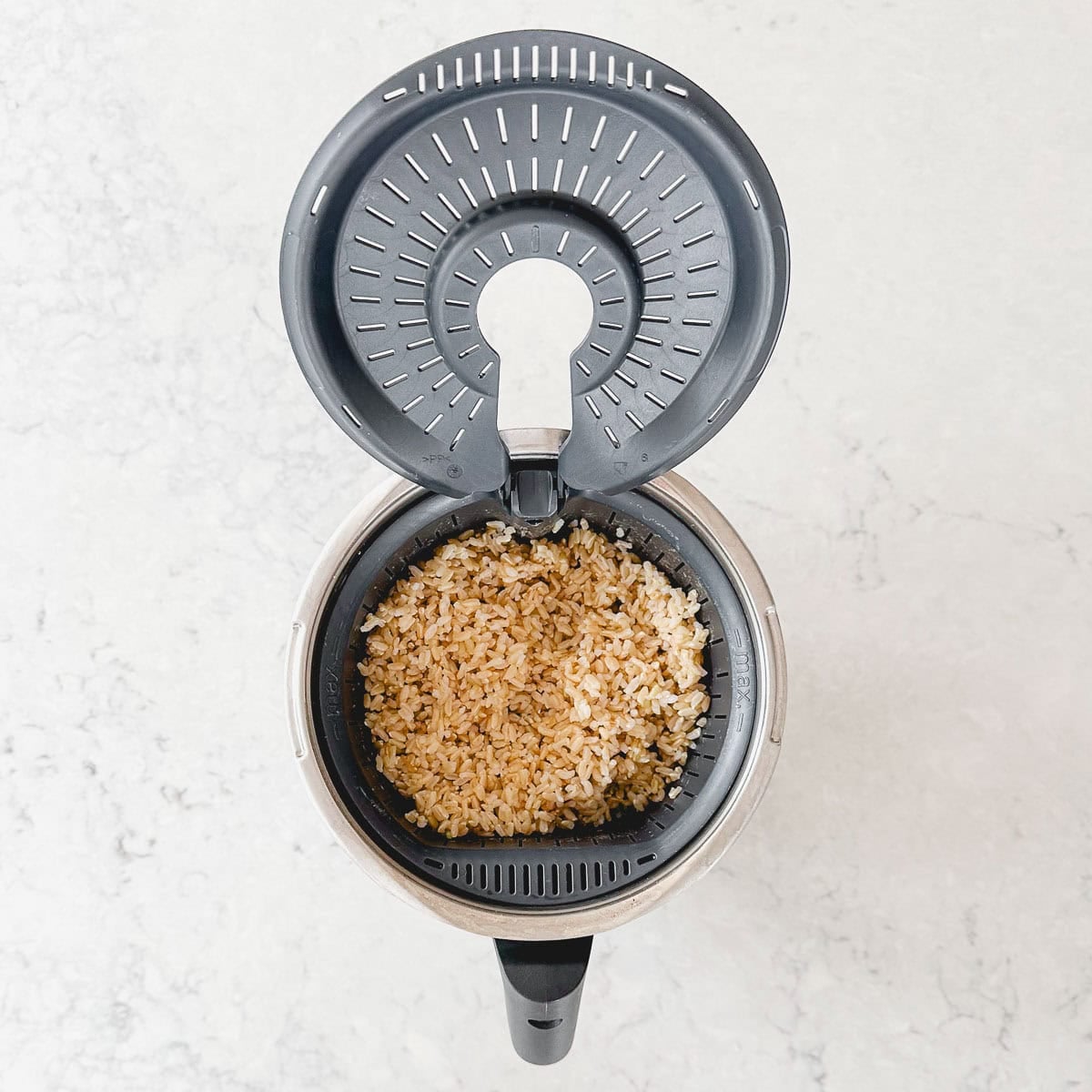 A top-down view of a Thermomix bowl with its lid open, showing cooked brown rice inside the inner basket on a light, marbled surface.
