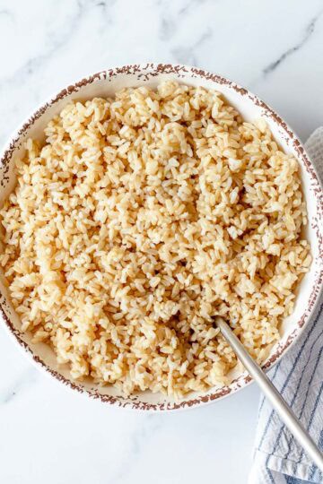 A bowl of cooked brown rice with a spoon inside, placed on a white marble surface next to a striped tea towel.