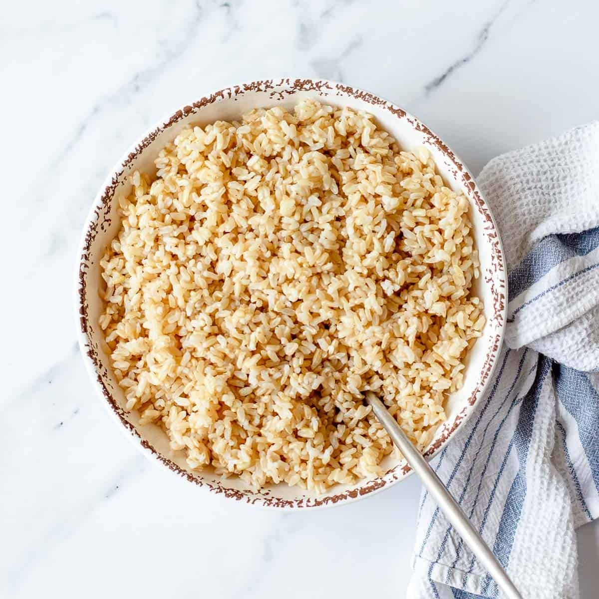 A bowl of cooked brown rice with a spoon inside, placed on a white marble surface next to a striped tea towel.