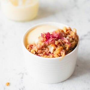 A white ramekin filled with rhubarb crumble topped with a crumble mixture and served with creamy custard, placed on a white marble surface.