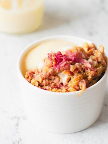 A white ramekin filled with rhubarb crumble topped with a crumble mixture and served with creamy custard, placed on a white marble surface.