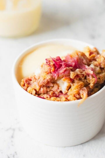 A white ramekin filled with rhubarb crumble topped with a crumble mixture and served with creamy custard, placed on a white marble surface.