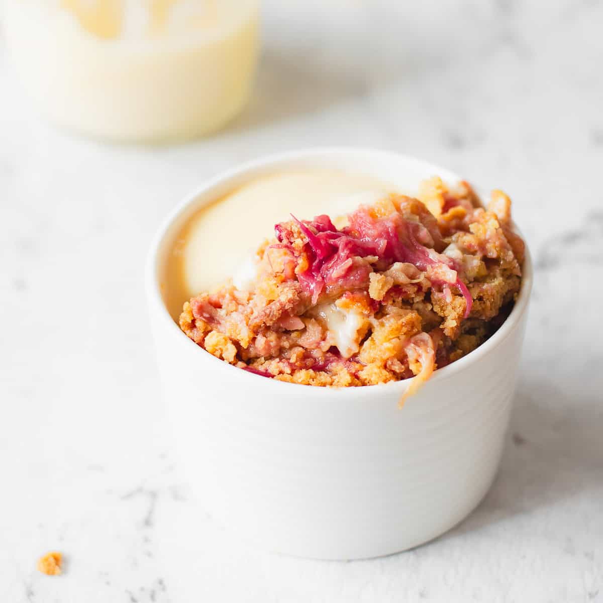 A white ramekin filled with rhubarb crumble topped with a crumble mixture and served with creamy custard, placed on a white marble surface.