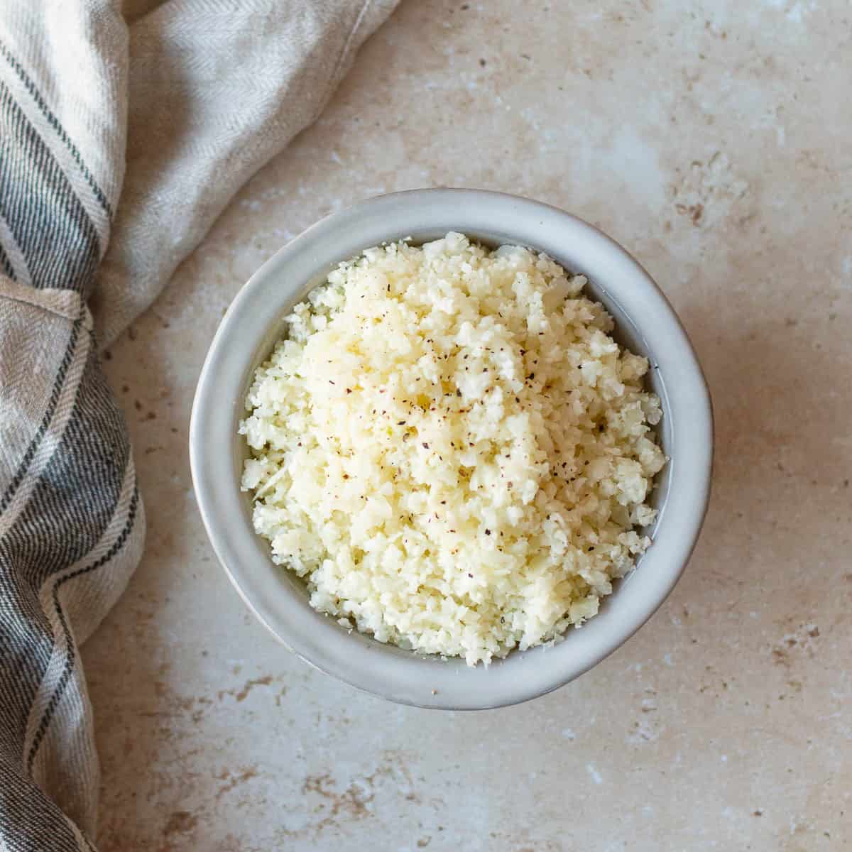 A top down view of a bowl of cauliflower rice, sitting on a marble benchtop with a tea towel to the left.