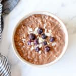 Chocolate Porridge in a white bowl with blueberries and coconut chips.
