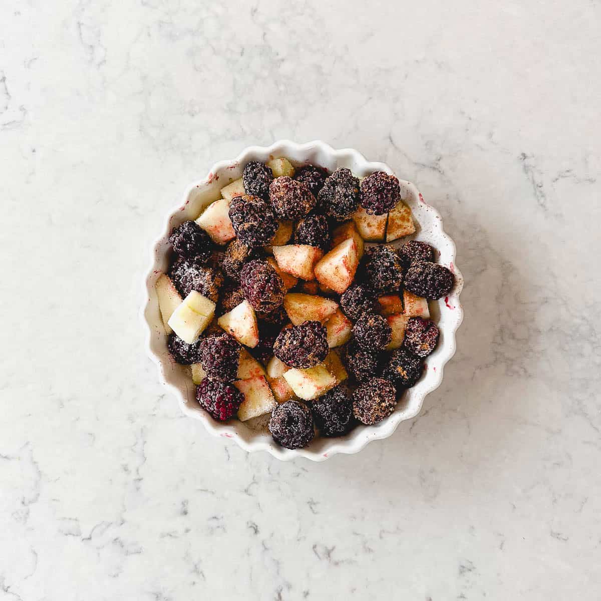 A white dish filled with chopped apples and blackberries, sprinkled with sugar and cinnamon, sits on a light marble surface.