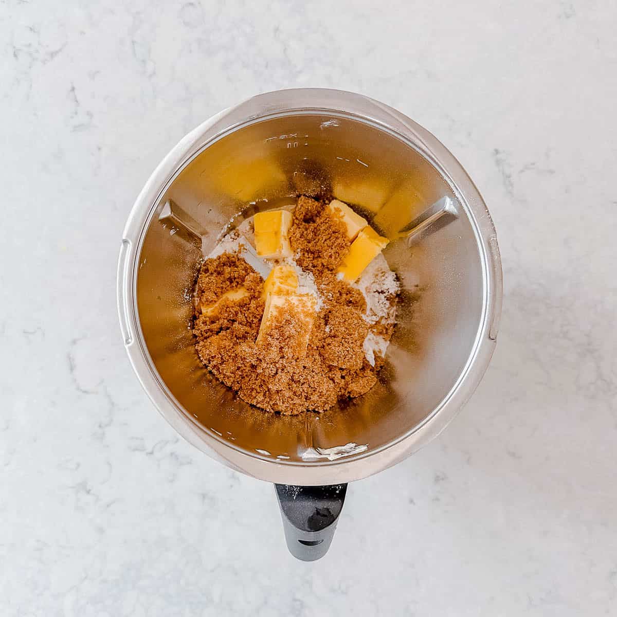 A top-down view of a metal mixing bowl containing chunks of butter and a pile of brown sugar on a light-coloured worktop.