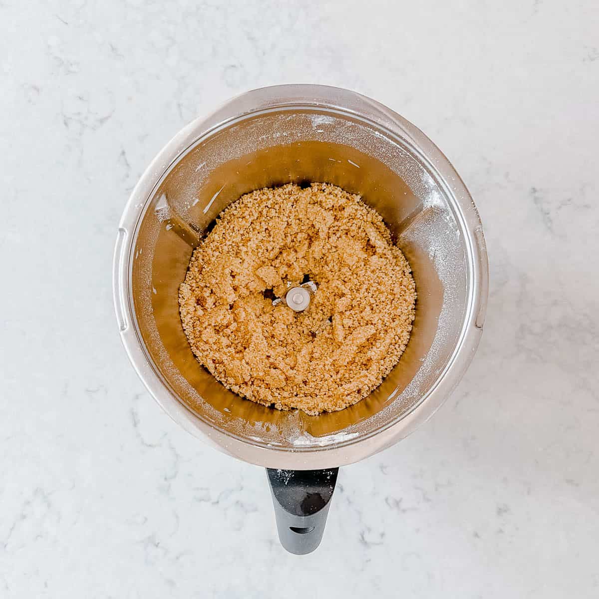 Overhead view of a food processor containing light brown, crumbly mixture, on a white marble surface.