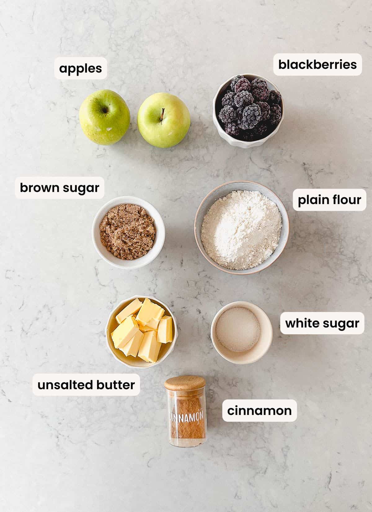 Flat lay of labelled baking ingredients on a marble surface: two green apples, a bowl of blackberries, bowls of brown sugar, plain flour, and white sugar, cubes of unsalted butter, and a jar of cinnamon.