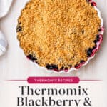 A baked blackberry and apple crumble in a round dish sits on a light surface next to a striped cloth. Text on the image reads, Thermomix Blackberry & Apple Crumble.