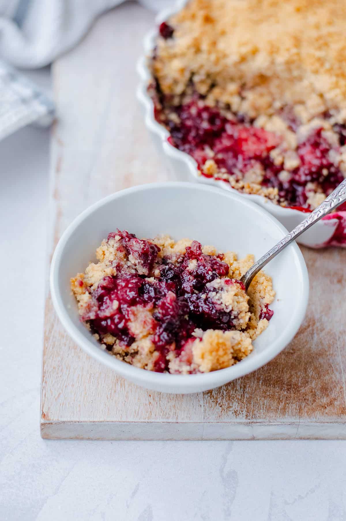 A white bowl filled with berry crumble pudding sits on a wooden board, with a spoon inside. In the background, a larger dish of the crumble, partially served, is visible.