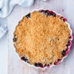 A round baking dish filled with berry crumble, showing a golden, crumbly topping with bits of fruit filling bubbling at the edges, set on a light wooden surface with a striped tea towel nearby.