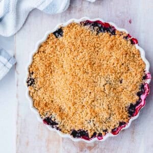 A round baking dish filled with berry crumble, showing a golden, crumbly topping with bits of fruit filling bubbling at the edges, set on a light wooden surface with a striped tea towel nearby.