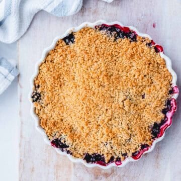 A round baking dish filled with berry crumble, showing a golden, crumbly topping with bits of fruit filling bubbling at the edges, set on a light wooden surface with a striped tea towel nearby.