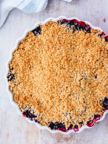 A round baking dish filled with berry crumble, showing a golden, crumbly topping with bits of fruit filling bubbling at the edges, set on a light wooden surface with a striped tea towel nearby.