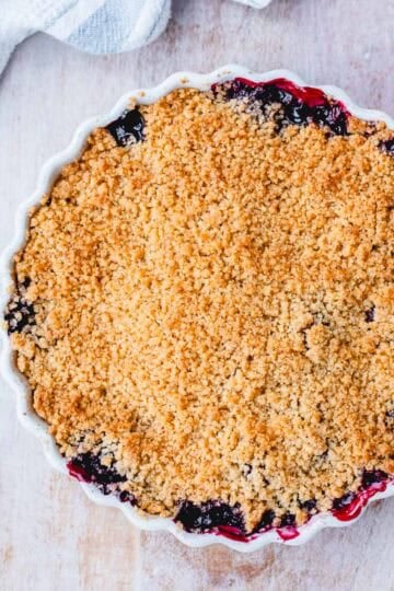 A round baking dish filled with berry crumble, showing a golden, crumbly topping with bits of fruit filling bubbling at the edges, set on a light wooden surface with a striped tea towel nearby.