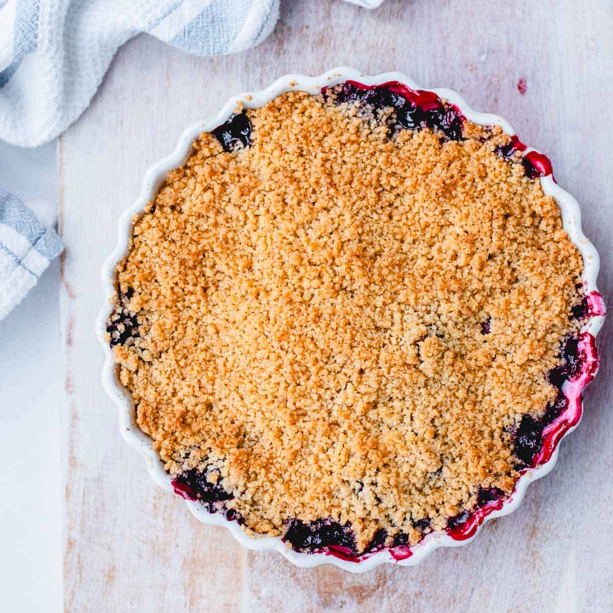 A round baking dish filled with berry crumble, showing a golden, crumbly topping with bits of fruit filling bubbling at the edges, set on a light wooden surface with a striped tea towel nearby.