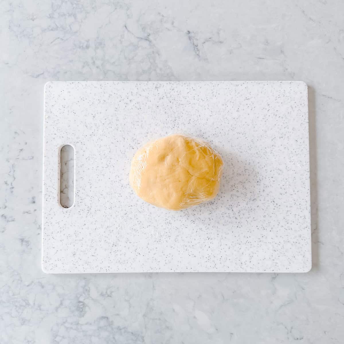 Pastry shaped into a disc shape, covered in plastic wrap and sitting on a white chopping board.
