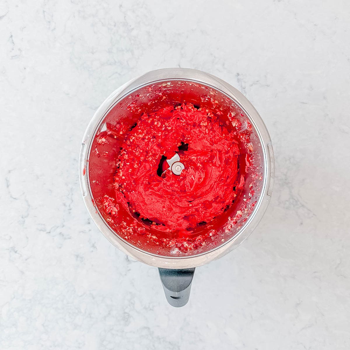 A food processor bowl filled with bright red, puréed mixture with a visible metal blade in the centre, set on a light, marbled worktop.