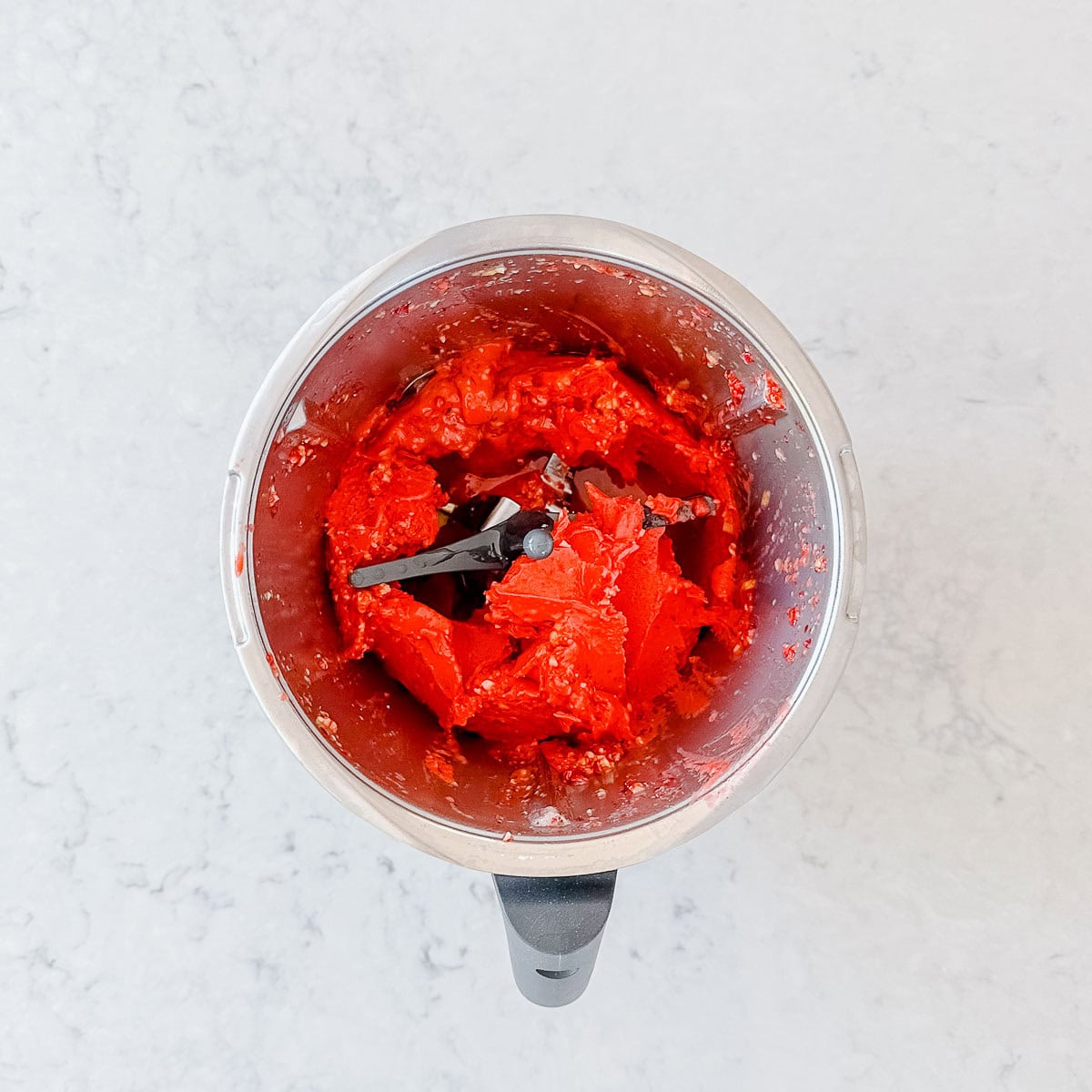 A food processor with bright red blended mixture inside, viewed from above, on a light-coloured countertop.