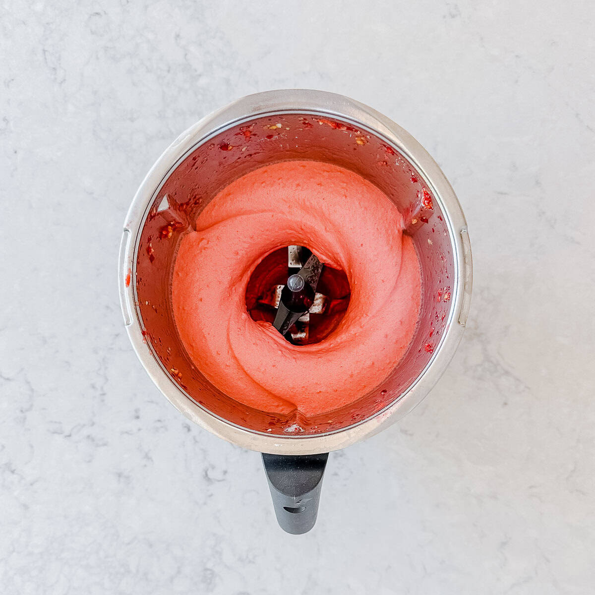 A blender viewed from above contains a smooth, pink mixture with a creamy texture, swirled around the blender blades against a light, speckled worktop background.