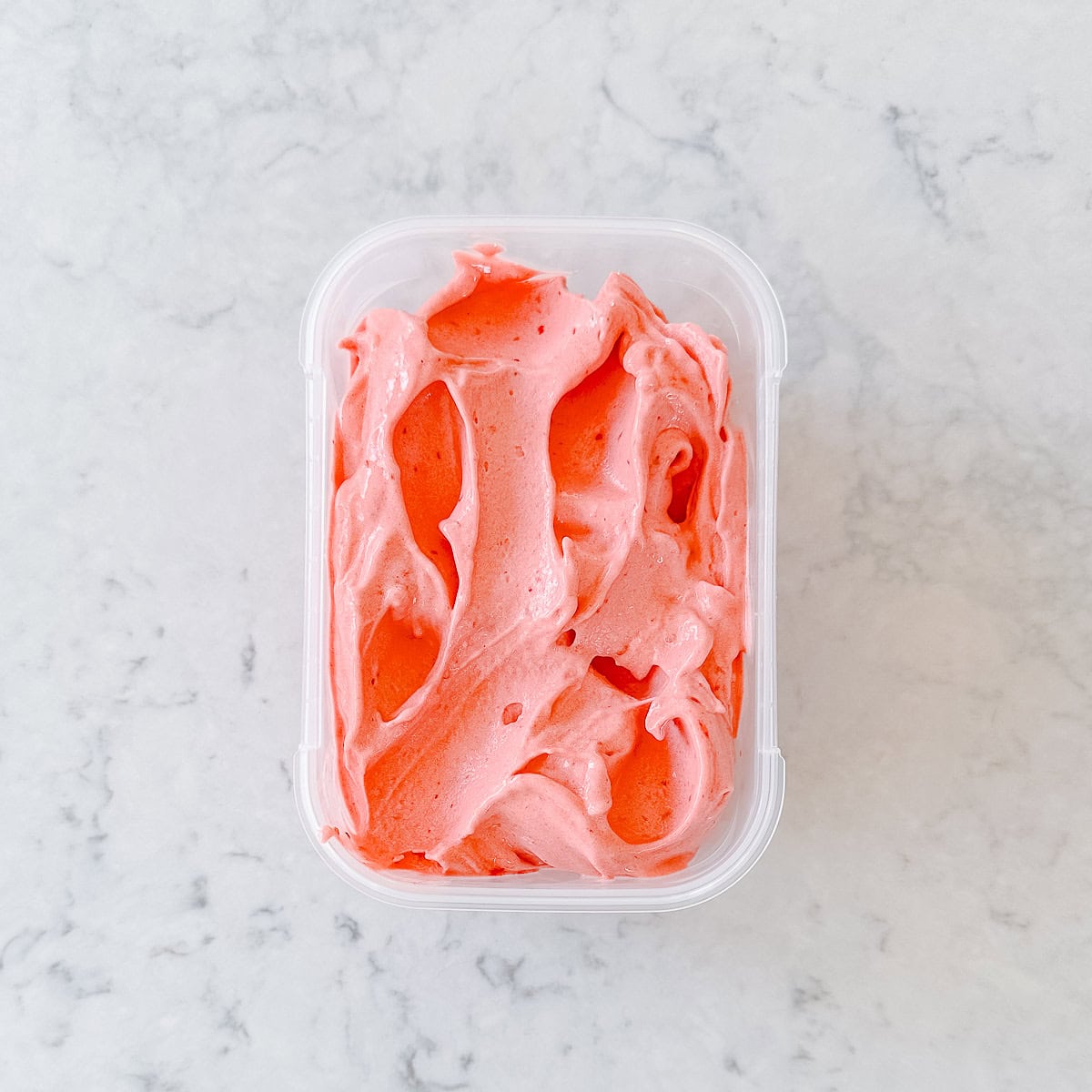 A rectangular plastic container filled with smooth, swirled pink sorbet sits on a white marble surface.