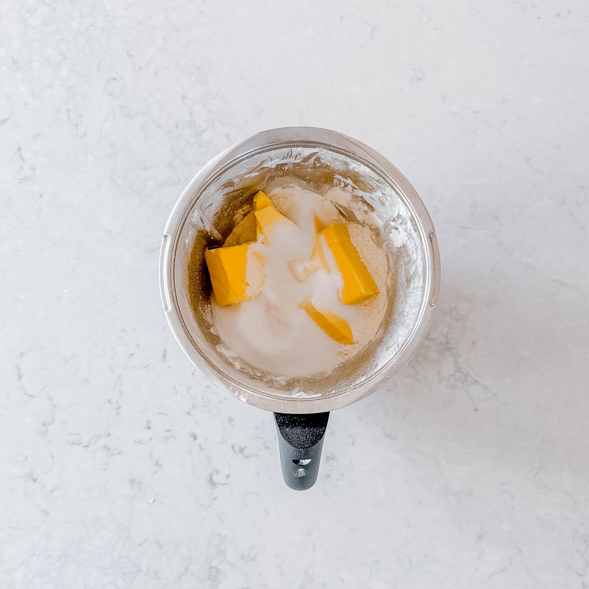 A mixing bowl contains chunks of butter and sugar, partially blended, on a white marble countertop.