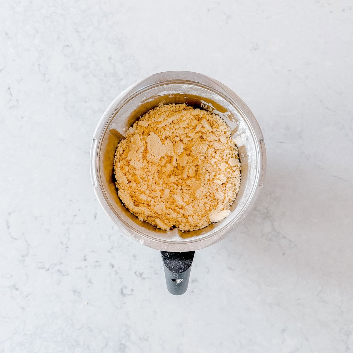 A Thermomix bowl filled with shortbread mixture sits on a light marble countertop.