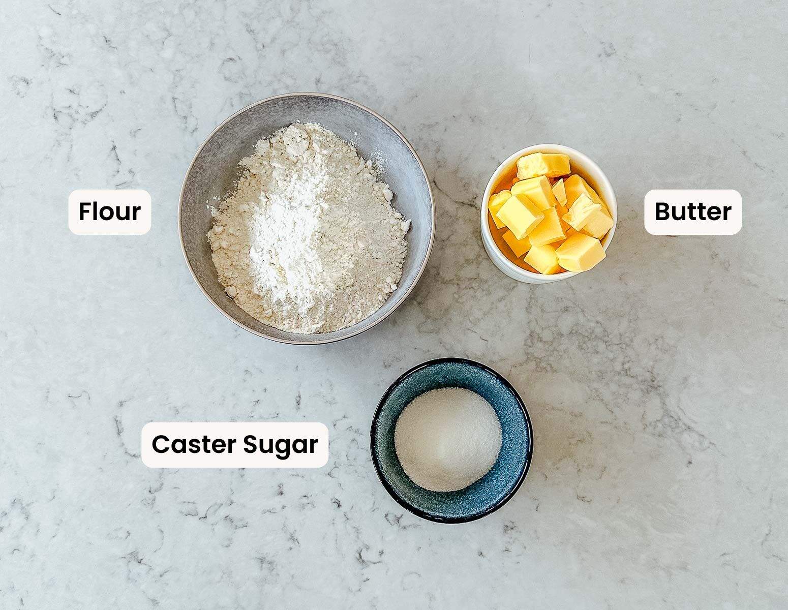 Overhead image of a bowl of flour, butter and sugar with a label next to each one, on a marble benchtop.