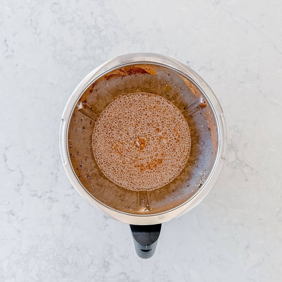 A top-down view of a blender filled with a light brown, frothy liquid, sitting on a white marbled worktop.
