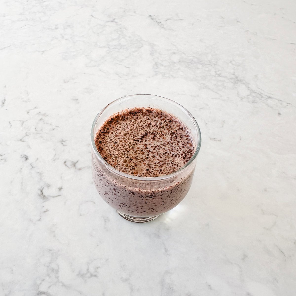 A glass filled with a chocolate chia pudding sits on a light-coloured, marbled surface.