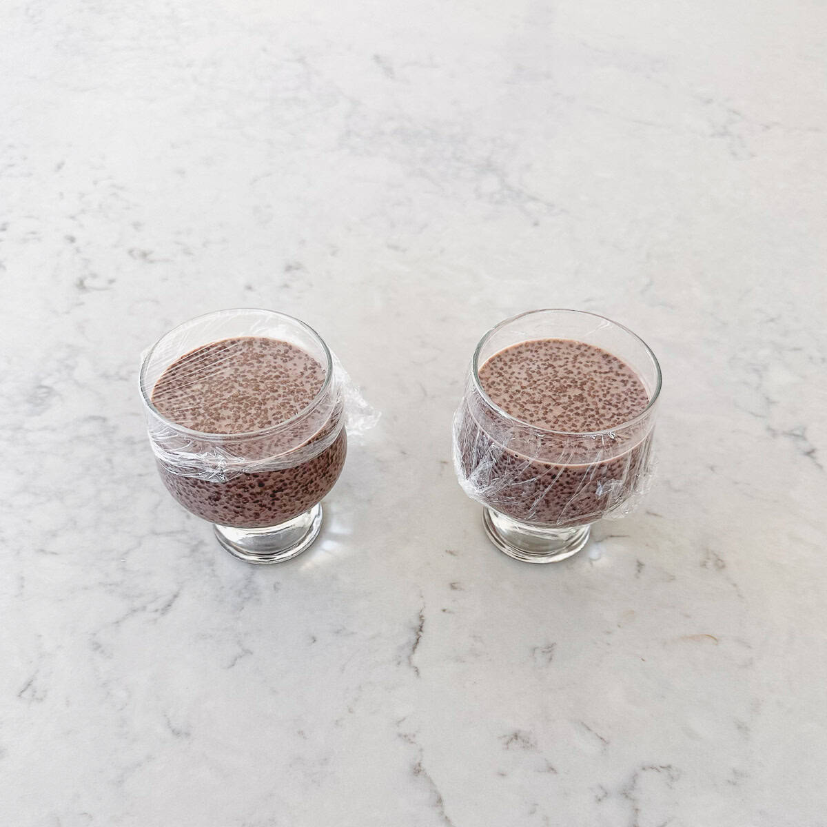 Two small glasses filled with chocolate chia pudding are covered with cling film and placed on a light-coloured, marble worktop.