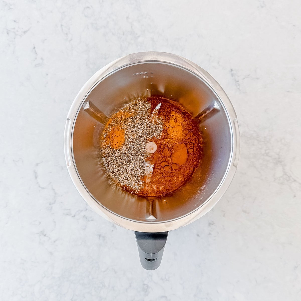 A Thermomix bowl seen from above contains ground spices and honey on a white marble surface, ready to be mixed.