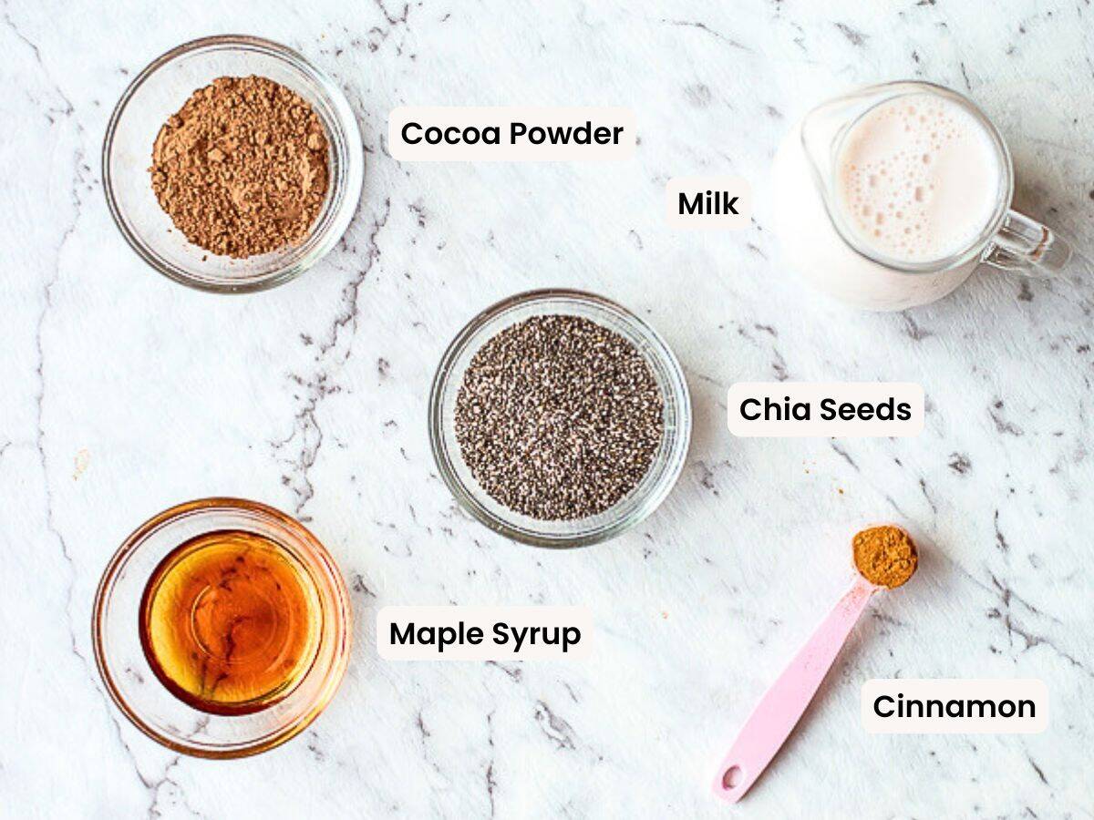 Five ingredients for a recipe are displayed on a white marble surface: cocoa powder in a bowl, a glass of milk, chia seeds in a bowl, maple syrup in a small bowl, and cinnamon on a pink spoon. Each is labelled.
