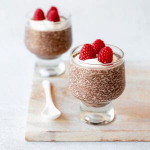 Two glass tumblers filled with chocolate chia pudding, topped with whipped cream and fresh raspberries, are placed on a light wooden board. A white ceramic spoon rests next to the front tumbler.