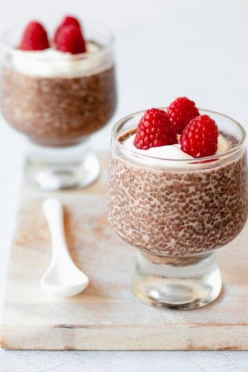 Two glass tumblers filled with chocolate chia pudding, topped with whipped cream and fresh raspberries, are placed on a light wooden board. A white ceramic spoon rests next to the front tumbler.