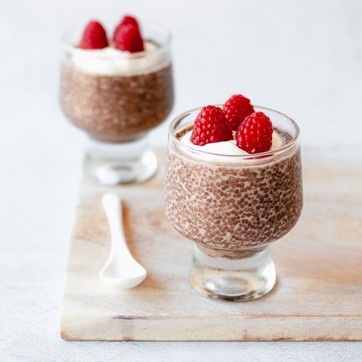 Two glass tumblers filled with chocolate chia pudding, topped with whipped cream and fresh raspberries, are placed on a light wooden board. A white ceramic spoon rests next to the front tumbler.