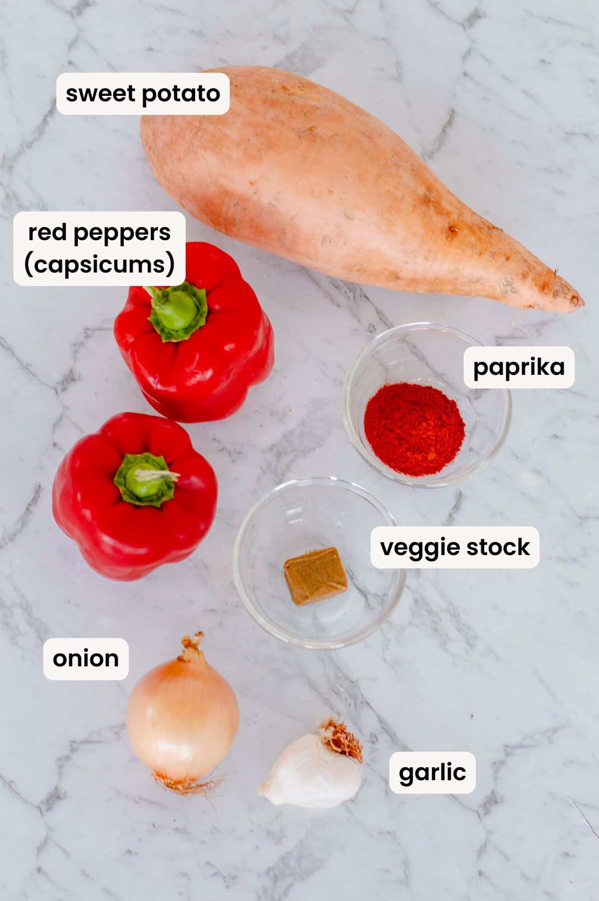 A sweet potato, two red peppers, a small bowl of paprika, a vegetable stock cube in a bowl, a yellow onion, and a garlic bulb, all labelled, arranged on a marble surface.