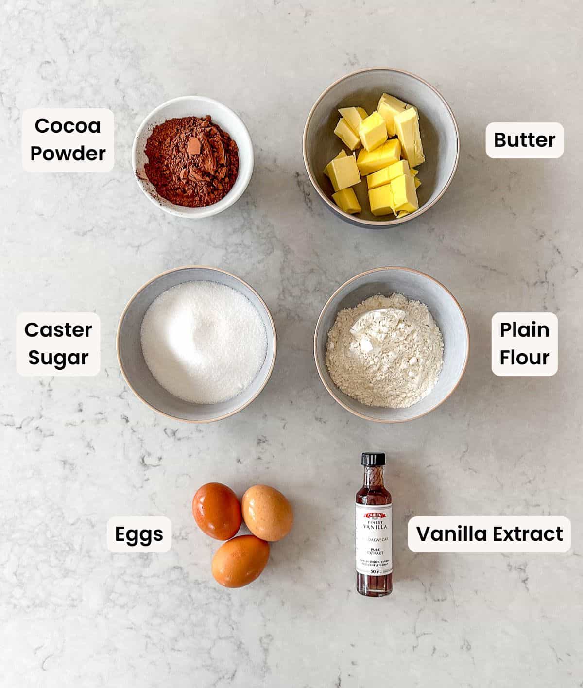 Six bowls and containers on a light surface hold plain flour, caster sugar, cubed butter, cocoa powder, peanut butter, and three eggs, each labelled with its ingredient name.