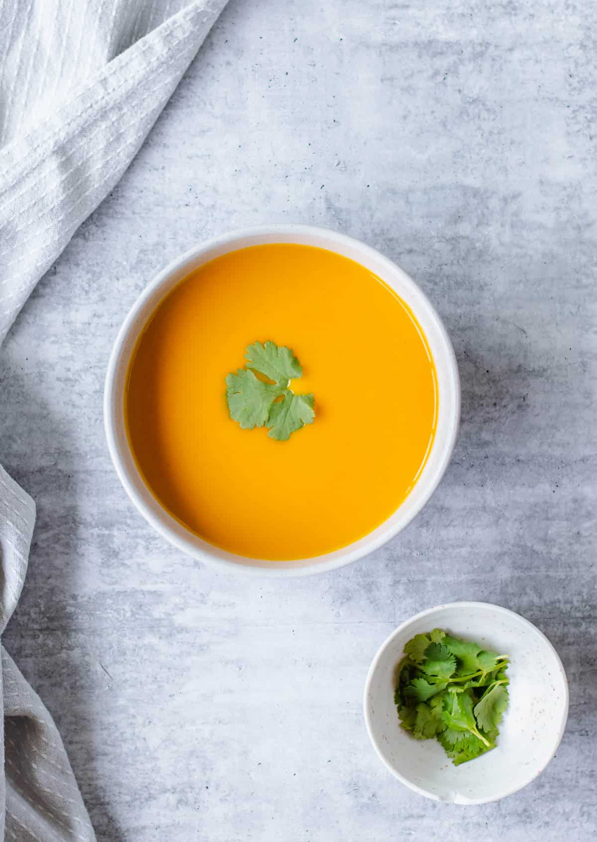 Overhead image of carrot and coriander soup on a a grey background with a small bowl of fresh coriander.